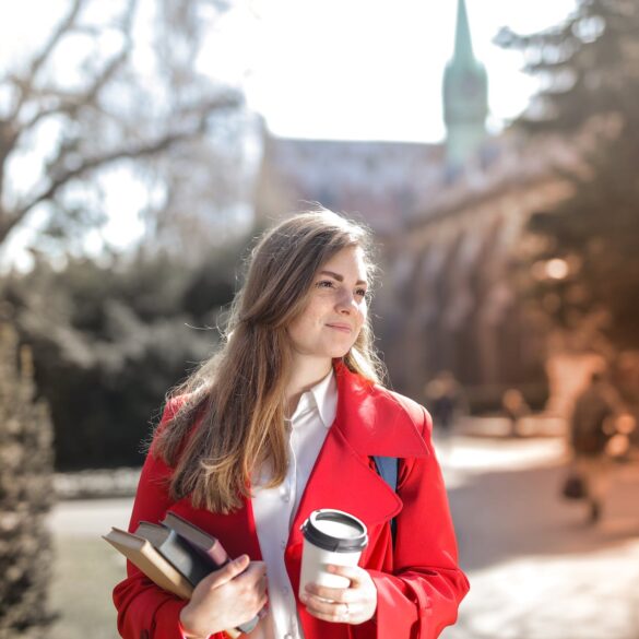 woman in red coat holding notebooks and coffee cup
