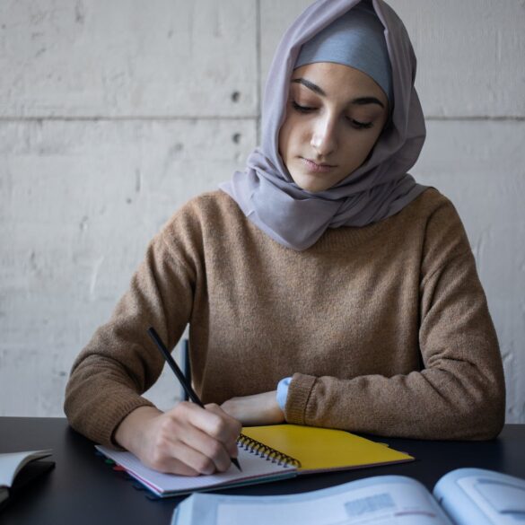diligent ethnic woman in hijab taking notes in workbook during lesson