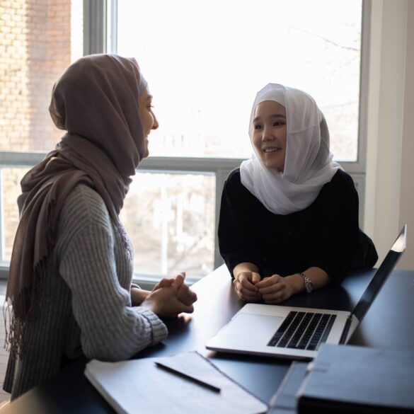 cheerful diverse women in hijabs communicating while sitting at table with laptop